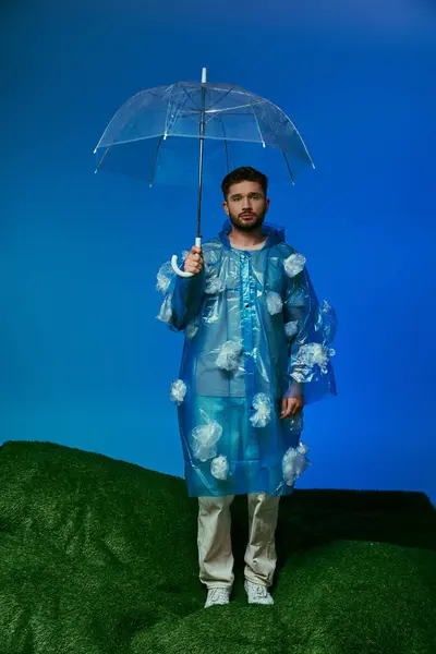 A man in a plastic raincoat holds a clear umbrella on a green hill in front of a blue background. — Stock Photo