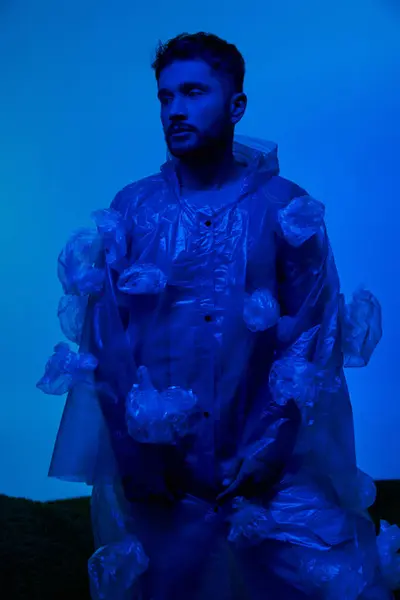 Un homme se tient sur un fond bleu, enveloppé dans un manteau de sacs en plastique. — Photo de stock