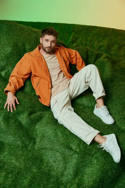 A man sits on a green carpet, wearing an orange jacket and white pants. — Stock Photo