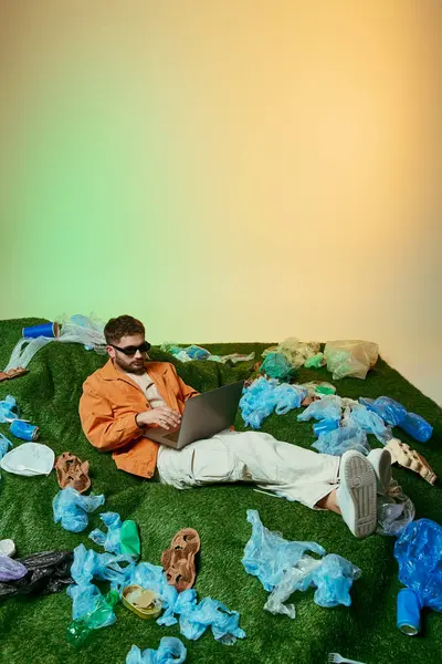 A man lies on a patch of artificial grass surrounded by plastic waste, working on a laptop. — Stock Photo
