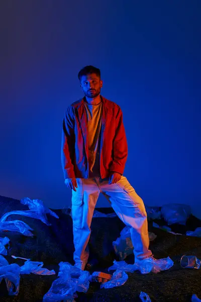 A young man stands on a pile of plastic waste, illuminated by vibrant blue and red lights. — Stock Photo