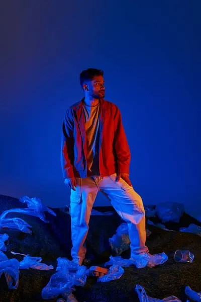 A man stands amidst a sea of plastic waste, bathed in vibrant blue and red light. — Stock Photo