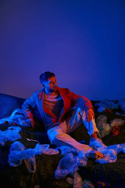 Un hombre se sienta entre un mar de bolsas de plástico, bañado en vibrante luz azul y roja. — Stock Photo