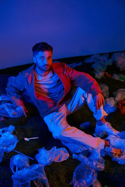 A man sits surrounded by plastic waste, illuminated by colorful lights. — Stock Photo