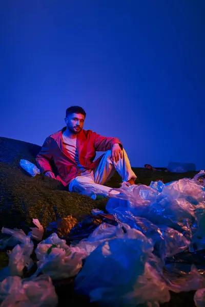 A young man sits amongst a sea of discarded plastic bags, illuminated by a blue and purple light. — Stock Photo
