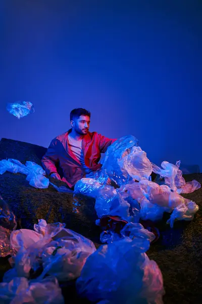 A young man sits amidst a sea of plastic bags, illuminated by a blue and red light. — Stock Photo