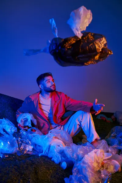 A man surrounded by plastic waste in a neon-lit landscape, reaching out towards a floating bag. — Stockfoto