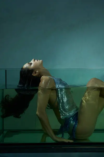 A young woman with wet brunette hair floats in an aquarium, her body submerged in the water, while wearing a shimmering top and skirt. — Stock Photo