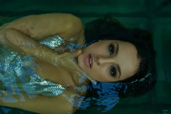 A young woman with wet hair floats in an aquarium, her eyes gazing upwards through the shimmering water. — Stock Photo