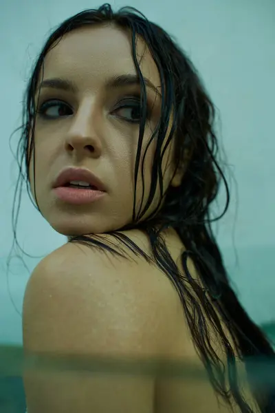 A young woman with wet, dark hair gazes over her shoulder while submerged in a pool of clear water. — Stock Photo