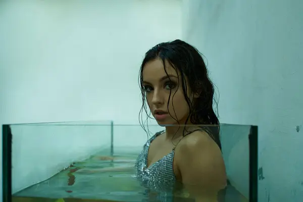 A beautiful young woman with wet hair, wearing a shimmering top, emerges from a glass tank filled with water. — Stock Photo