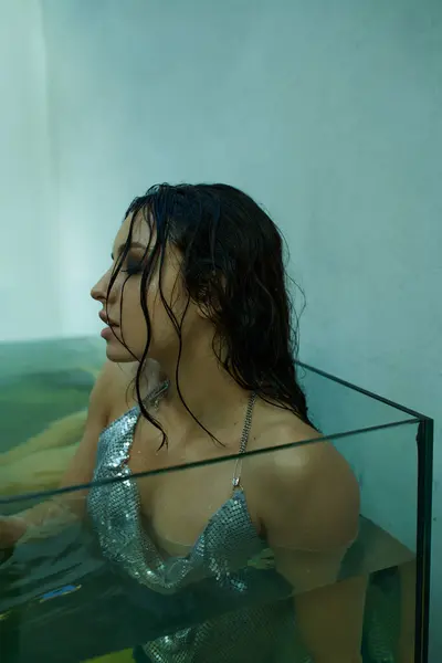 A young woman with wet, dark hair, poses in a glass aquarium, wearing a shimmering silver top. — Stock Photo