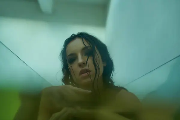 A young woman with wet hair emerges from the water, her gaze piercing through the glass. — Stock Photo
