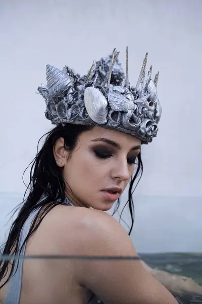 A young woman with long wet hair and a silver seashell crown looks down at the water, submerged in a tank. — Stock Photo