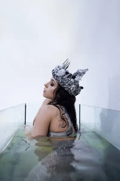 A young woman with long dark hair, wearing a seashell crown, is submerged in water in a glass tank, with a thoughtful expression. — Stock Photo