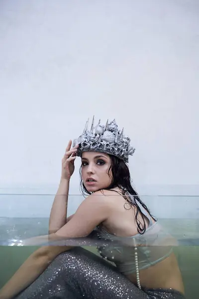 A young woman wearing a silver seashell crown sits in a glass tank filled with water, her mermaid tail shimmering beneath the surface. — Stock Photo
