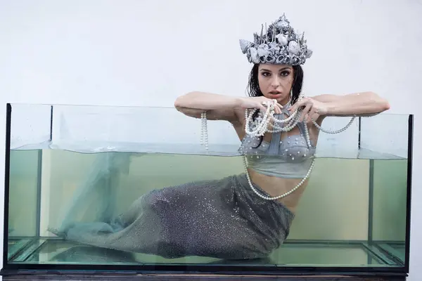 A mermaid, wearing a silver crown and pearl necklace, leans against the edge of a glass tank, her tail disappearing into the water. — Stock Photo