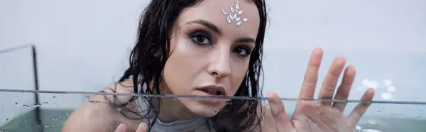 A woman with dark hair and a jeweled forehead, looks out from an aquarium, with water rippling around her. — Stock Photo