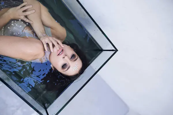 A young woman with dark hair floats in a clear tank, her eyes closed and her hands resting on her chest. — Stock Photo