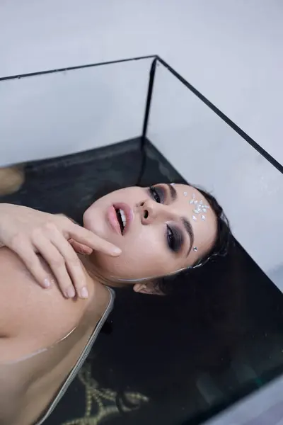 A young woman with dark hair and striking makeup floats serenely within a glass aquarium. — Stock Photo