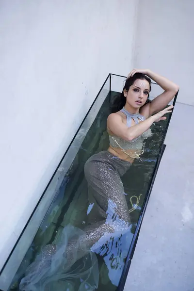 A young woman with brunette hair poses as a mermaid in a rectangular aquarium, her shimmering tail catching the light. — Stock Photo