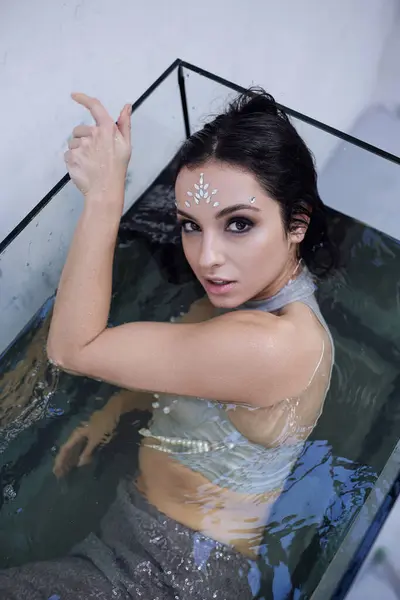 A young woman with dark hair and glittering face paint poses in an aquarium, looking up at the camera. — Stock Photo