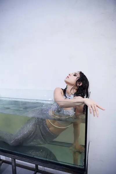 A young woman dressed as a mermaid poses in a glass aquarium, her hair wet and cascading around her shoulders. — Stock Photo