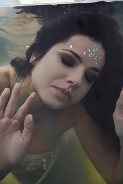 A young woman with dark hair and a jeweled forehead floats beneath the surface of an aquarium, her eyes closed in a dreamlike state. — Stock Photo