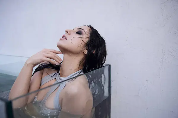 A beautiful woman with long brunette hair floats in a glass tank filled with water, her eyes closed in contemplation. — Stock Photo