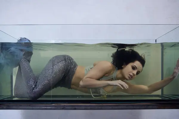 A young woman in a mermaid costume swims underwater in a large glass tank, her expression wistful. — Stock Photo