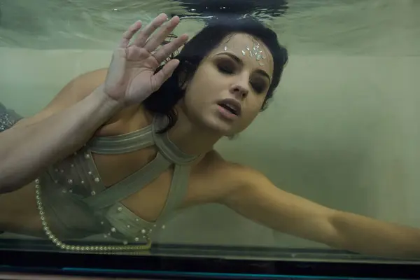 A young woman, adorned with shimmering makeup, swims in an aquarium, her eyes fixed on the viewer. — Stock Photo