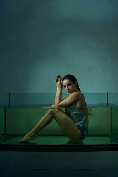A young woman with wet hair sits in a clear aquarium, gazing intently at the viewer. — Stock Photo