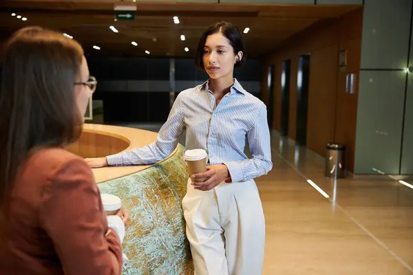 Two colleagues chat in an office lobby. One woman is holding a cup of coffee. — Stock Photo