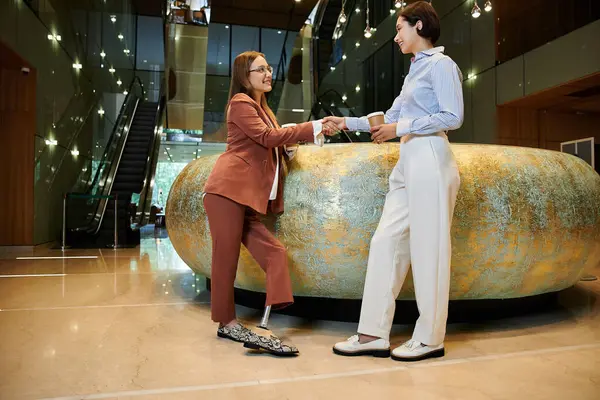 Two coworkers, a woman with a prosthetic leg and her colleague, chat near an office escalator over coffee in a modern lobby. — Stock Photo