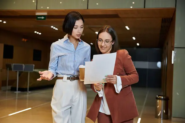 Two women in professional attire converse and review documents in a modern office lobby. — Stock Photo
