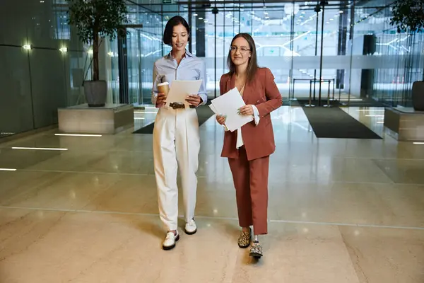 Two female colleagues, one with a prosthetic leg, walk and chat while holding papers in an office lobby. — Stock Photo