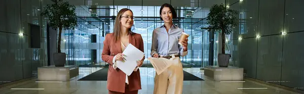 Two colleagues chat in a modern office lobby, one holding a cup of coffee and the other carrying paperwork. — Stock Photo