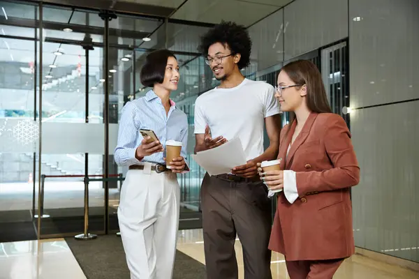 Three colleagues, stand in a glass-walled lobby, discussing work over coffee. — Stock Photo