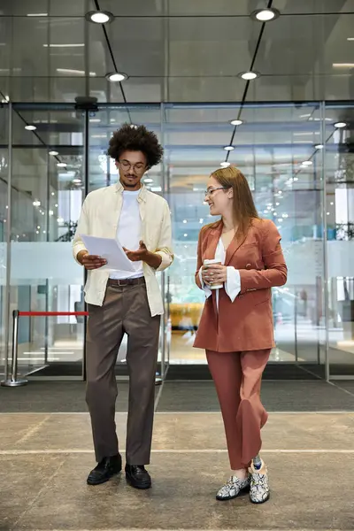 Ein Mann und eine Frau plaudern lässig in einer Büro-Lobby, die Frau hält eine Kaffeetasse in der Hand und der Mann prüft Dokumente. — Stockfoto