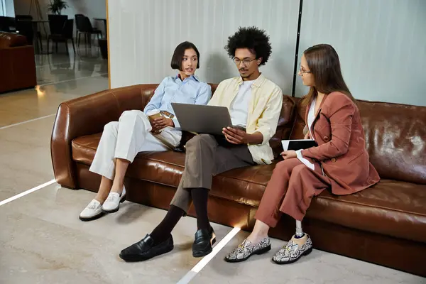 Three colleagues, including a woman with a prosthetic leg, sit on a brown leather sofa in an office lobby, looking at a laptop. — Stock Photo