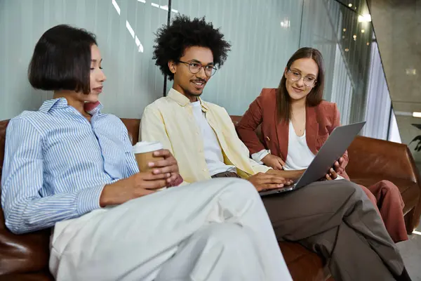 Three colleagues sit on a couch in an office lobby, chatting and looking at a laptop. — Stock Photo