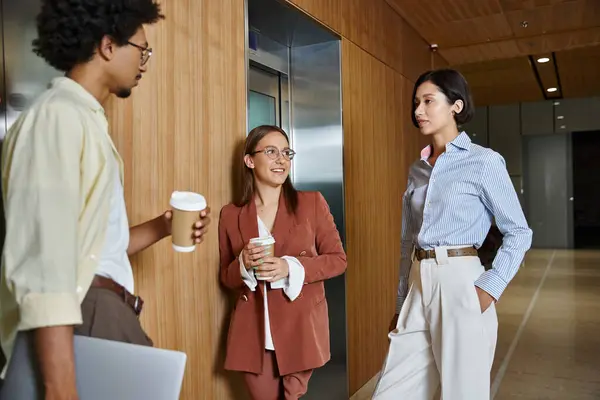 Three colleagues stand in a modern office hallway, chatting and holding coffee cups. — Stock Photo