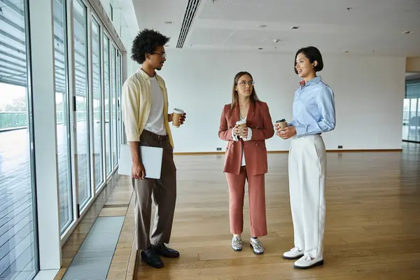 Three colleagues in a modern office chat while holding coffee cups, showcasing diversity and inclusion. — Stock Photo