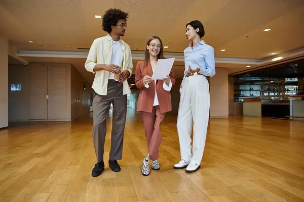 Three colleagues, diverse in ethnicity and background, stand in a modern office, engaging in conversation. — Stock Photo