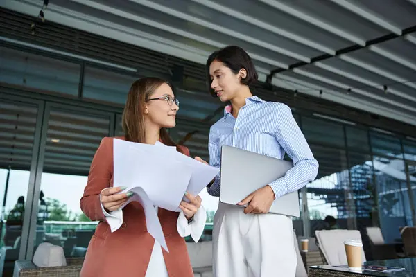 Two colleagues, one holding a laptop and the other reviewing documents, chat on a modern office rooftop terrace. — Stock Photo