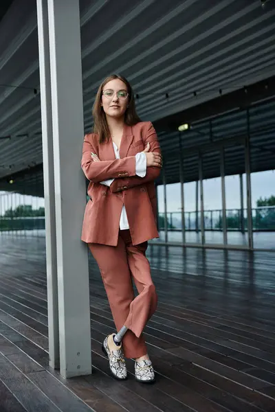 Eine Frau im rosafarbenen Anzug steht selbstbewusst auf einer modernen Dachterrasse und lehnt an einer Säule. Sie trägt ein weißes Hemd und eine Brille. — Stockfoto