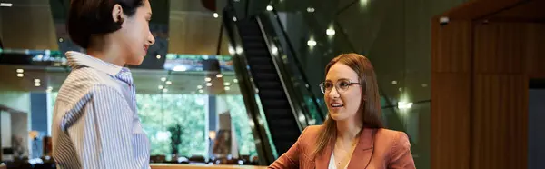 Two coworkers chat and smile in an office lobby. One wears a striped shirt, the other a blazer and glasses. — Stock Photo