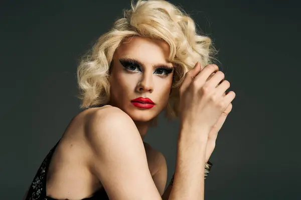 Una drag queen con capelli biondi e trucco, indossa un lungo abito nero, posa con un'espressione seria su uno sfondo grigio. — Foto stock