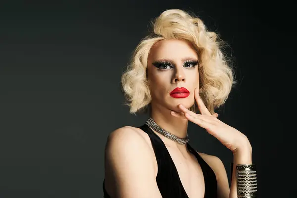 Uma drag queen com maquiagem impecável e uma peruca loira glamourosa posa em um vestido preto contra um pano de fundo cinza. — Fotografia de Stock