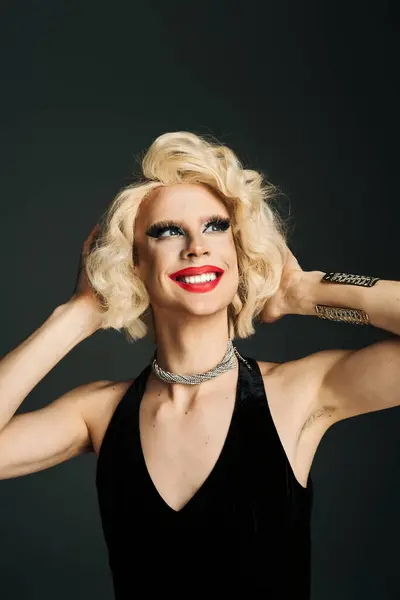 A drag queen with blonde hair and makeup smiles and poses in a black dress against a gray background. — Stock Photo